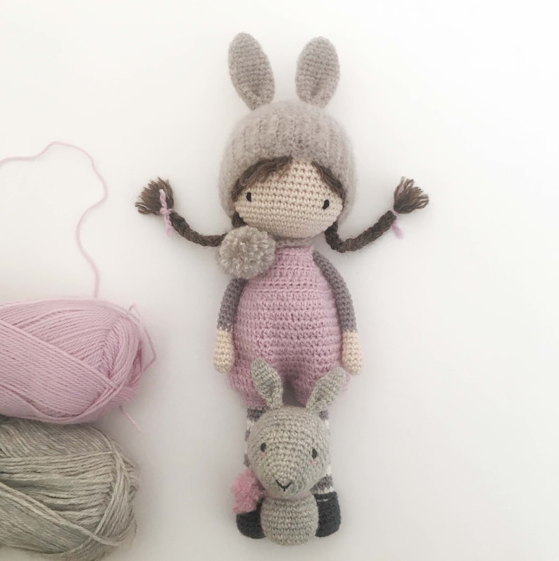 Pode incluir: Boneca de croch&ecirc; feita &agrave; m&atilde;o com um chap&eacute;u de coelho e um brinquedo de coelho a condizer. A boneca usa um vestido rosa e tem tran&ccedil;as castanhas com detalhes rosa. Meadas de l&atilde; cinza e rosa est&atilde;o no fundo.