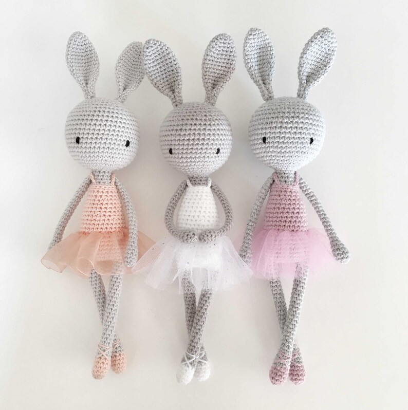 Puede incluir: Tres mu&ntilde;ecas de conejo de crochet vestidas con tut&uacute;s de bailarina. Los conejos son grises con ojos negros y llevan tut&uacute;s rosa, blanco y rosa claro.