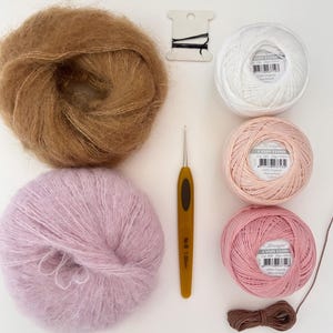 May include: An overhead shot of various craft supplies, including balls of yarn in brown, white, pink, and lavender. A crochet hook, black thread, and a brown ribbon are also present. The yarn balls are labeled "Candy Floss".