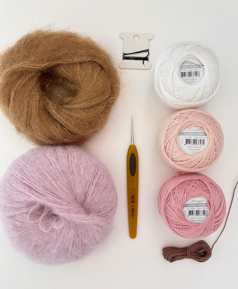 May include: An overhead shot of various craft supplies, including balls of yarn in brown, white, pink, and lavender. A crochet hook, black thread, and a brown ribbon are also present. The yarn balls are labeled "Candy Floss".