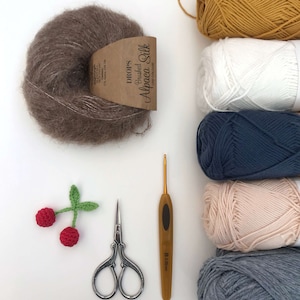 Puede incluir: Una colecci&oacute;n de ovillos de lana en varios colores, incluyendo marr&oacute;n, amarillo, blanco, azul, rosa y gris. Tambi&eacute;n hay un gancho de ganchillo, un par de tijeras y una peque&ntilde;a cereza de ganchillo con un tallo verde.