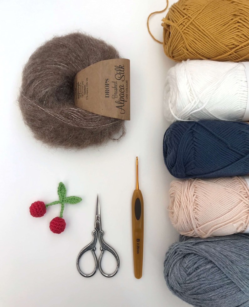 Puede incluir: Una colecci&oacute;n de ovillos de lana en varios colores, incluyendo marr&oacute;n, amarillo, blanco, azul, rosa y gris. Tambi&eacute;n hay un gancho de ganchillo, un par de tijeras y una peque&ntilde;a cereza de ganchillo con un tallo verde.