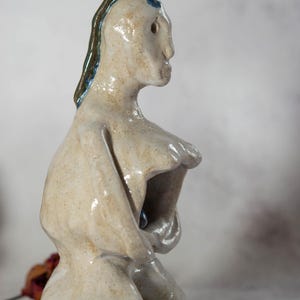 May include: A ceramic sculpture of a figure with a light beige glaze. The figure has a stylized form with a prominent chest and a head with dark brown and blue accents. The sculpture is on a reflective surface.