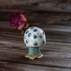May include: A ceramic mushroom figurine with blue and white polka dots and a green base. The mushroom is sitting on a wooden surface.