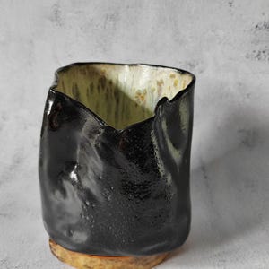 May include: A black ceramic vase with a textured, uneven surface and a light brown base. The vase has a unique, organic shape and a glossy finish.