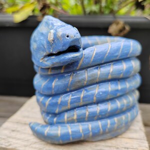 May include: A blue ceramic snake sculpture with white stripes coiled into a spiral shape. The sculpture is sitting on a wooden surface.