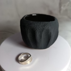 May include: A black, textured ceramic bowl with a smooth, dark interior and a silver ring. The bowl has a rough, organic exterior. The ring has a simple, modern design. The items are displayed on a white surface.