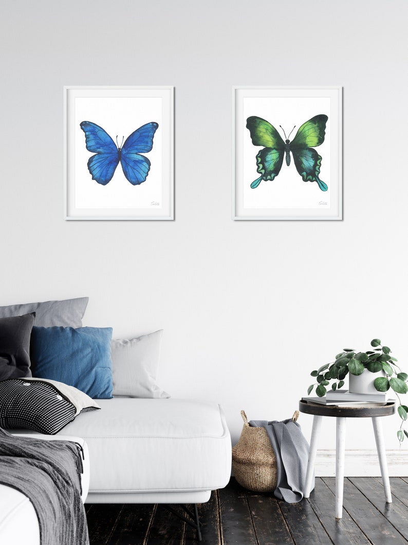 Puede incluir: Dos pinturas de acuarela enmarcadas de mariposas. La mariposa de la izquierda es azul y la mariposa de la derecha es verde. Ambas mariposas est&aacute;n en vuelo.
