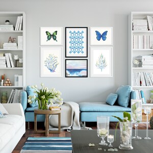 Puede incluir: Un interior de sala de estar con un sof&aacute; azul claro, una mesa de caf&eacute; blanca y estantes blancos con libros. La pared presenta una galer&iacute;a de obras de arte enmarcadas, que incluyen un patr&oacute;n geom&eacute;trico azul y blanco, una pintura de acuarela de un paisaje marino y dos ilustraciones de mariposas.