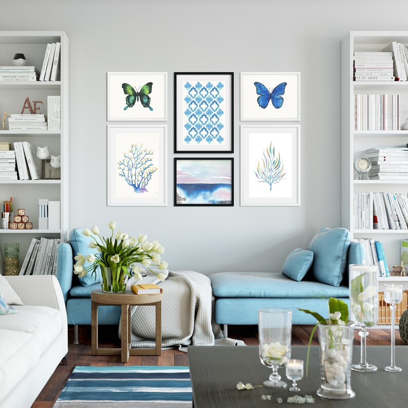 Puede incluir: Un interior de sala de estar con un sof&aacute; azul claro, una mesa de caf&eacute; blanca y estantes blancos con libros. La pared presenta una galer&iacute;a de obras de arte enmarcadas, que incluyen un patr&oacute;n geom&eacute;trico azul y blanco, una pintura de acuarela de un paisaje marino y dos ilustraciones de mariposas.