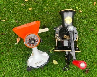 Puede incluir: Dos molinillos de comida manuales vintage. Uno es negro con detalles dorados y dice "The New Coffee Mill". El otro es blanco con la parte superior naranja y dice "Spong Nao's".