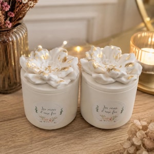 Peut inclure: Deux contenants ronds blanc cassé avec des couvercles en forme de fleur. Les couvercles sont blancs avec des accents dorés. Chaque contenant porte une étiquette avec le texte "Les rêves d'une fée" et des illustrations florales. Sur une surface en bois.