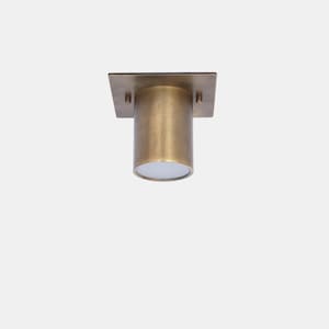 May include: A brass-coloured ceiling light fitting with a square base and a cylindrical light housing. The light fitting has a simple, modern design and is suitable for various interior styles. The light housing is approximately 10 cm in length.