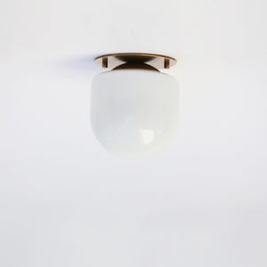 May include: A modern ceiling light fixture with a white, rounded glass shade and a bronze-colored metal base. The light is designed to be mounted flush against the ceiling. The fixture has a simple, minimalist design.