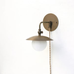 May include: A brass wall sconce with a round shade and a white globe light bulb. The sconce has a cord with a plug.