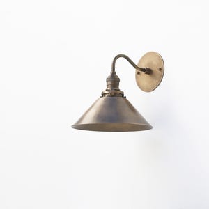 May include: A brass wall sconce with a cone-shaped shade. The sconce has a simple design and a vintage aesthetic.