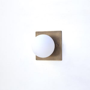 May include: A modern wall sconce featuring a square, aged brass-toned backplate and a spherical, frosted white glass shade. The fixture is designed for indoor lighting and has a minimalist aesthetic.