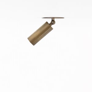 May include: A cylindrical brass spotlight fixture featuring a circular mounting plate and an adjustable swivel joint. The metal surface has a brushed finish, and the light head is angled downward, connected to the ceiling mount by a small hinge.