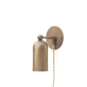May include: A brass wall-mounted light fixture with a bell-shaped shade and a round wall plate. The light fixture has a cord with a braided fabric covering.