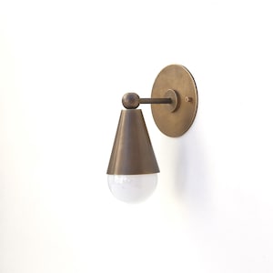 May include: A brass wall sconce featuring a conical shade and a circular backplate. The shade is a dark brass colour, and the bulb is a white globe shape. The sconce is mounted on a white wall.