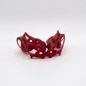 May include: A dark red, abstract bracelet with an organic, porous design. The bracelet features multiple openings and a flowing, sculptural form. The material appears to be a translucent, possibly resin-based, material. The bracelet is displayed against a white background.
