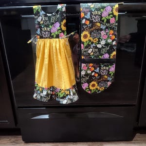 Op de afbeelding: Een gele keukendoek en ovenwant set. De handdoek heeft een zwarte bloemen top met de tekst "You can grow your own way" en een gele rok. De ovenwant heeft een zwart bloemenpatroon met zonnebloemen en de tekst "Be happy, your own way."