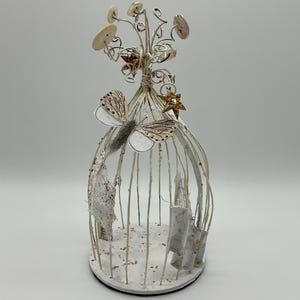 Pode incluir: Uma gaiola decorativa branca com enfeites de borboletas e estrelas. A gaiola apresenta um arranjo floral no topo e detalhes em papel no interior. A gaiola é feita de arame e assenta numa base redonda.