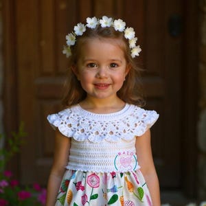 Könnte beinhalten: Ein Kleid mit weißem Häkeloberteil und einem Rock mit Blumenmuster in Rosa, Orange, Blau und Grün. Das Kleid hat einen geschwungenen Ausschnitt und ein weißes Blumenkranz-Accessoire. Ein kleines kreisförmiges Logo mit dem Text MAIO ist zu sehen.