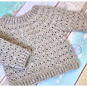 Pu&ograve; includere: Un maglione a maniche lunghe color beige chiaro realizzato con un motivo a uncinetto testurizzato, caratterizzato da scollo, polsini e orlo a coste. Un logo circolare con il testo MAIO CROCHET e un'icona di gomitolo &egrave; visibile in basso a destra.