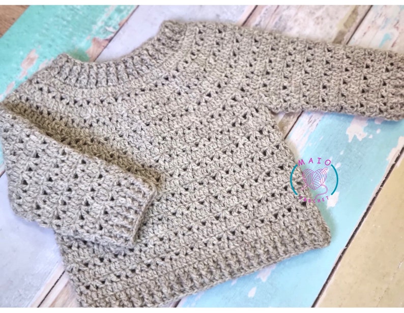 Pu&ograve; includere: Un maglione a maniche lunghe color beige chiaro realizzato con un motivo a uncinetto testurizzato, caratterizzato da scollo, polsini e orlo a coste. Un logo circolare con il testo MAIO CROCHET e un'icona di gomitolo &egrave; visibile in basso a destra.