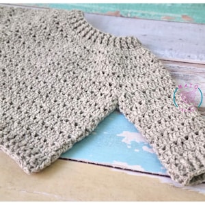 Pu&ograve; includere: Un maglione beige a maniche lunghe con un motivo a uncinetto testurizzato, scollo rotondo a coste e polsini a coste. Un logo circolare con il testo MAIO CROCHET &egrave; visibile sulla zona della manica destra di questo capo fatto a mano.
