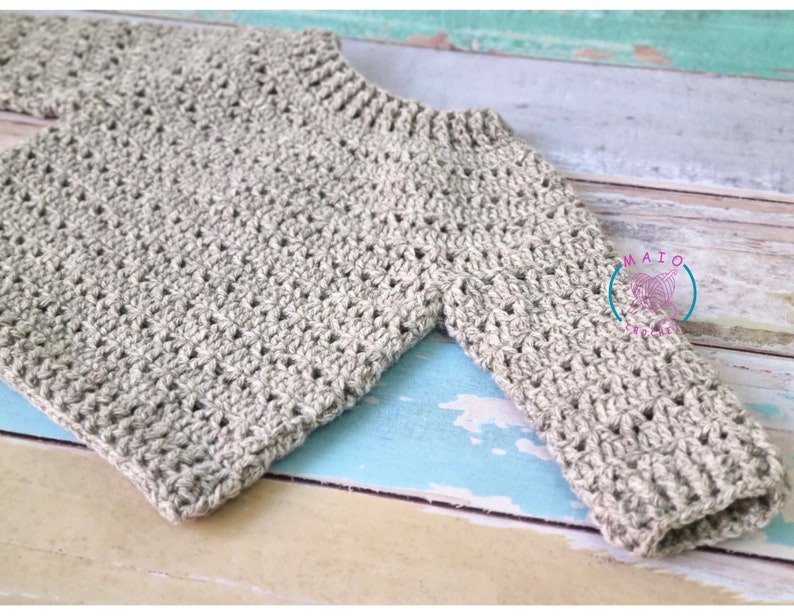 Pu&ograve; includere: Un maglione beige a maniche lunghe con un motivo a uncinetto testurizzato, scollo rotondo a coste e polsini a coste. Un logo circolare con il testo MAIO CROCHET &egrave; visibile sulla zona della manica destra di questo capo fatto a mano.