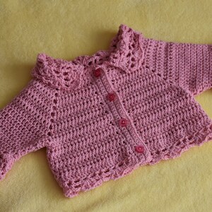 Può includere: Un cardigan rosa per neonato lavorato a uncinetto con colletto e polsini in pizzo. Il cardigan ha tre bottoni rosa sul davanti.