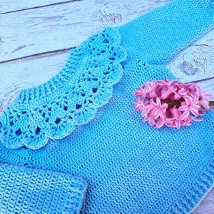 Può includere: Un maglione di lana blu chiaro con un grande colletto lavorato a uncinetto. Il maglione è appoggiato su una superficie di legno bianco con un fiore rosa.