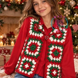 Può includere: Un gilet senza maniche all'uncinetto realizzato con quadrati granny rossi, bianchi e verdi. Il gilet presenta una chiusura con bottone a forma di cuore sul davanti ed è indossato sopra una camicia rossa a maniche lunghe e jeans blu.