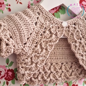 Könnte beinhalten: Hellbraune, gehäkelte Bolero-Jacke mit gewelltem Rand und einem einzelnen Knopfverschluss. Das kurzärmelige Kleidungsstück wird auf einem weißen Kleiderbügel vor einem floralen Hintergrund präsentiert. Die Häkelarbeit weist ein strukturiertes Muster auf.