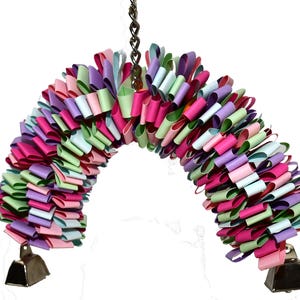 May include: A colorful, multi-layered, paper toy for birds, with pink, green, blue, and purple paper loops. The toy is hanging from a chain and has two silver bells at the bottom.