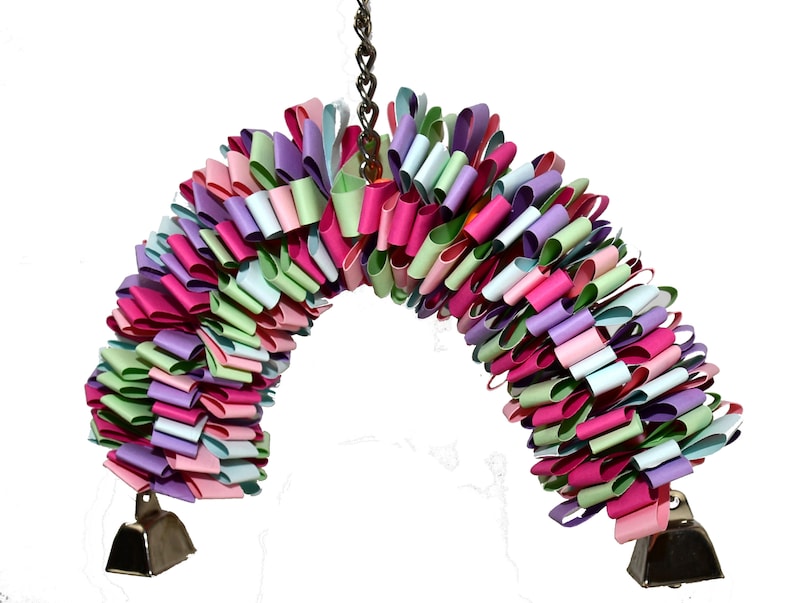 May include: A colorful, multi-layered, paper toy for birds, with pink, green, blue, and purple paper loops. The toy is hanging from a chain and has two silver bells at the bottom.