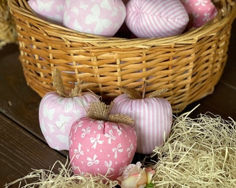 Puede incluir: Una cesta de mimbre llena de manzanas de tela rosa con varios patrones, incluyendo rayas, mariposas y lazos. Tres manzanas adicionales están delante de la cesta sobre una superficie de paja.