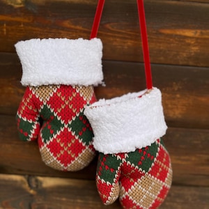Op de afbeelding: Een paar gebreide wanten met een ruitpatroon in rood, groen en beige, voorzien van witte boorden van imitatiebont. De wanten zijn verbonden door een dun rood fluwelen lint, waardoor ze samen als één decoratief item kunnen worden opgehangen.