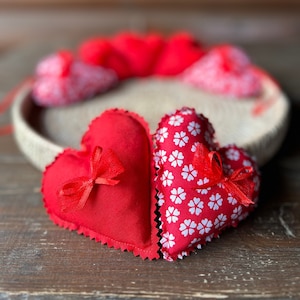 May include: Two red fabric hearts, one with a white floral pattern and the other solid red with a red bow. The hearts are sitting on a wooden surface.