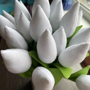 May include: A close-up of a bouquet of white fabric tulips with green fabric leaves. The tulip petals are a soft white, and the leaves are a bright green. The tulips are arranged in a small, square vase.