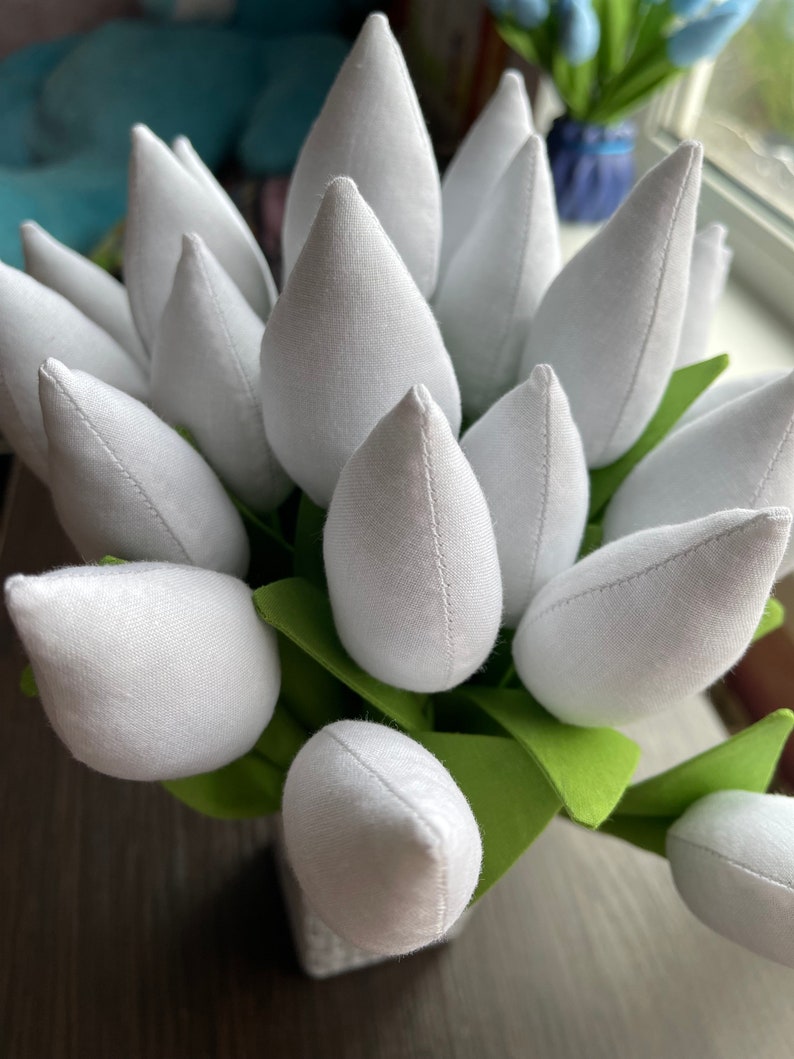 May include: A close-up of a bouquet of white fabric tulips with green fabric leaves. The tulip petals are a soft white, and the leaves are a bright green. The tulips are arranged in a small, square vase.