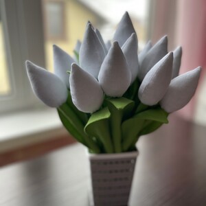 May include: A display of white fabric tulips in a square, silver vase. The tulip heads are a soft, light grey, with green leaves. The vase has a textured, dotted pattern. The arrangement is on a dark brown surface.