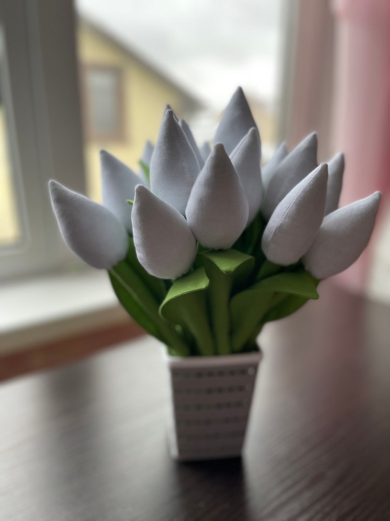 May include: A display of white fabric tulips in a square, silver vase. The tulip heads are a soft, light grey, with green leaves. The vase has a textured, dotted pattern. The arrangement is on a dark brown surface.