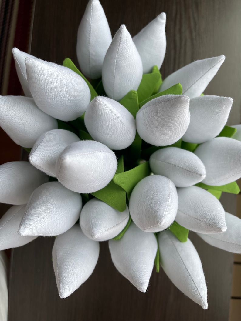 May include: A close-up of a bouquet of white fabric tulips with green fabric leaves. The tulips are tightly clustered, creating a full, textured appearance. The fabric has a slightly textured appearance, and the overall effect is soft and delicate.
