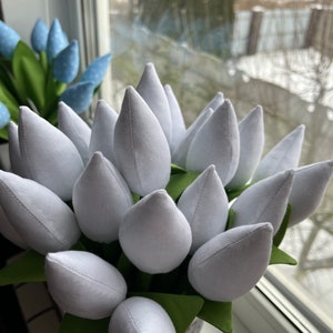 May include: A bunch of white fabric tulips with green fabric leaves, presented in a grey pot. A second bouquet of blue polka-dot tulips is visible in the background. The scene is set near a window.