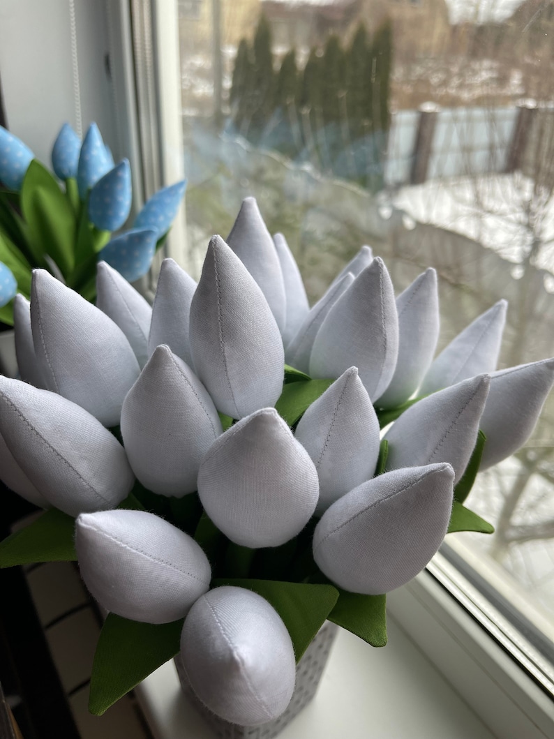 May include: A bunch of white fabric tulips with green fabric leaves, presented in a grey pot. A second bouquet of blue polka-dot tulips is visible in the background. The scene is set near a window.