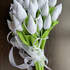 May include: A bouquet of white fabric tulips with green leaves and stems, tied with a white ribbon. The tulips are tightly clustered, creating a full and textured appearance. The background is a dark wood surface.