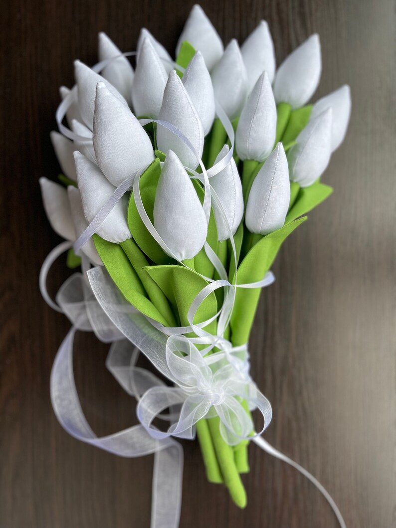 May include: A bouquet of white fabric tulips with green leaves and stems, tied with a white ribbon. The tulips are tightly clustered, creating a full and textured appearance. The background is a dark wood surface.